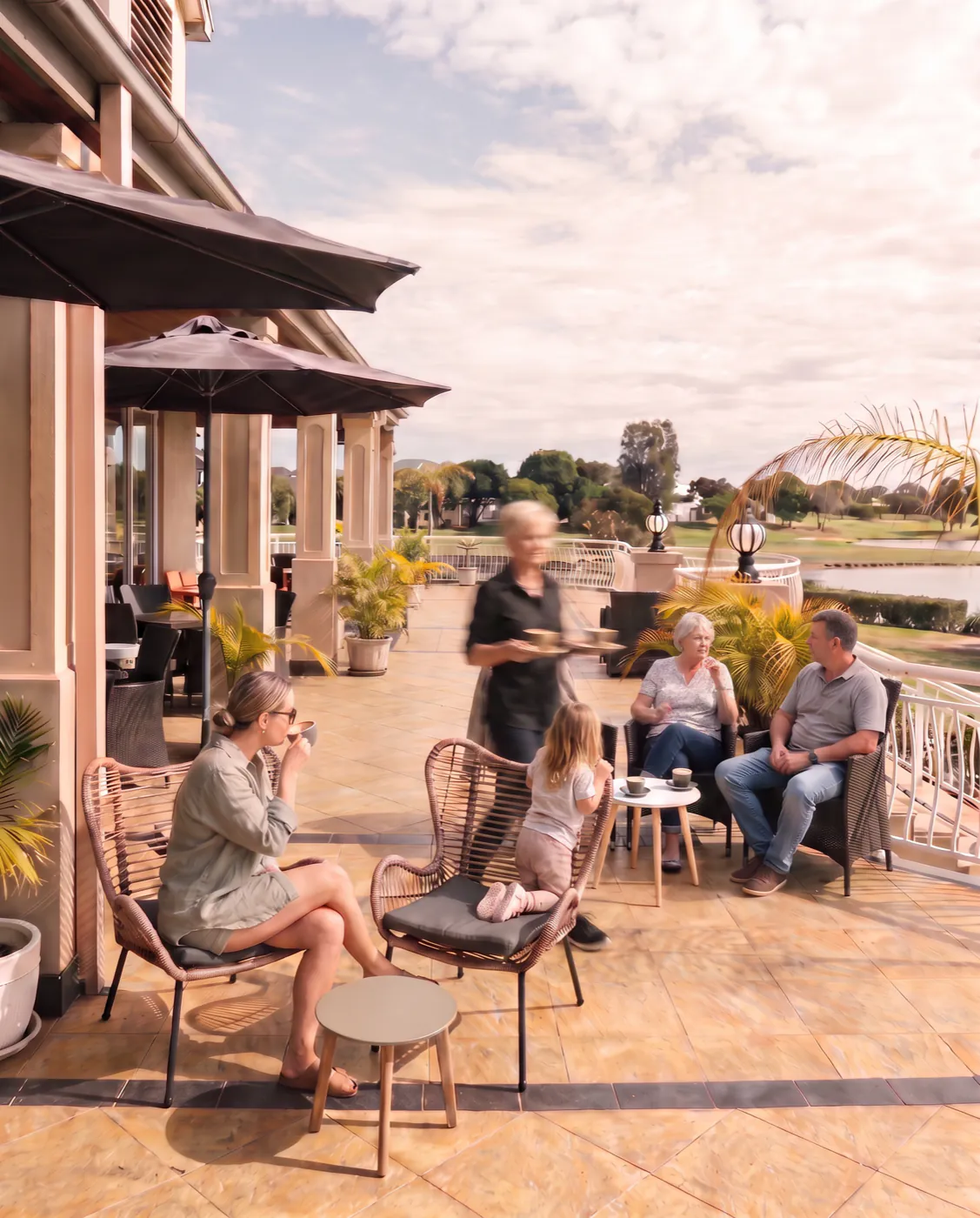 People over coffee on the clubhouse deck, with a member of staff carrying coffees past them and the fairway and the lake beyond the balustrade