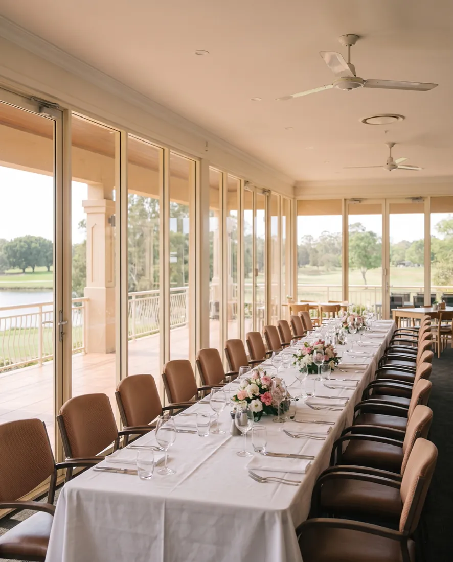A long table set with white linen, glassware and flowers in the clubhouse, along a wall of glass looking out over the lake and the fairway