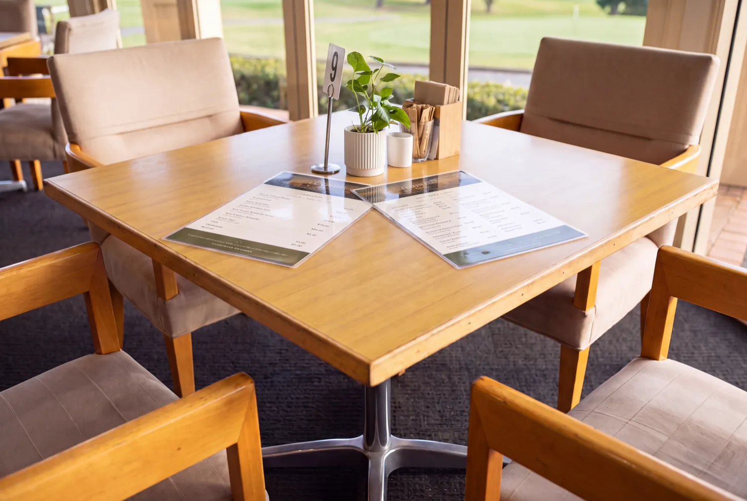 The breakfast menu and the all-day menu, laminated, on a table in the café beside the plant and napkins, with the golf course through the windows